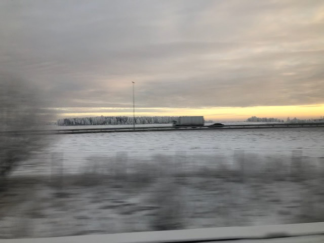 Grått vinterlandskap sett fra tog, med en lastebil og en personbil på motorveien midt i bildet.