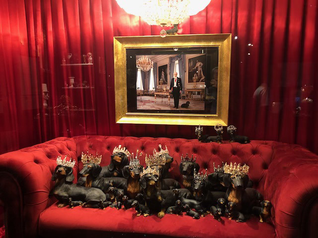 Fotografi av en stor gruppe dukker av sorte dachshunder, i livaktiv framtoning og forskjellige størrelser. Noen med krone på hodet. De sitter i en rød sofa omkranset av røde gardiner og i bakgrunnen er det et  fotografi av en adelig utseende mann med en annen dachs.