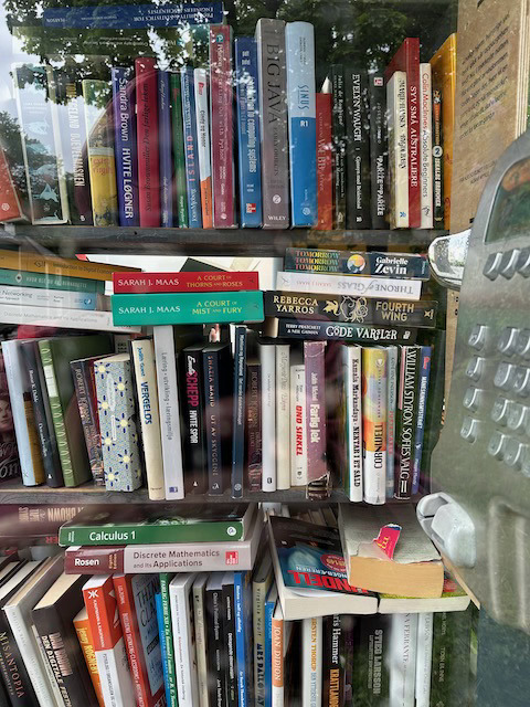 Bøker stablet i bokhylle sett gjennom vindu i gammel telefonkiosk med utsnitt av telefonen til høyre.