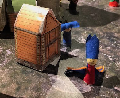 Fingerdukker av tøy og trekuler foran en papirmodell av en laftet trebu. Figurene har pavehatt og nonnehabitt.