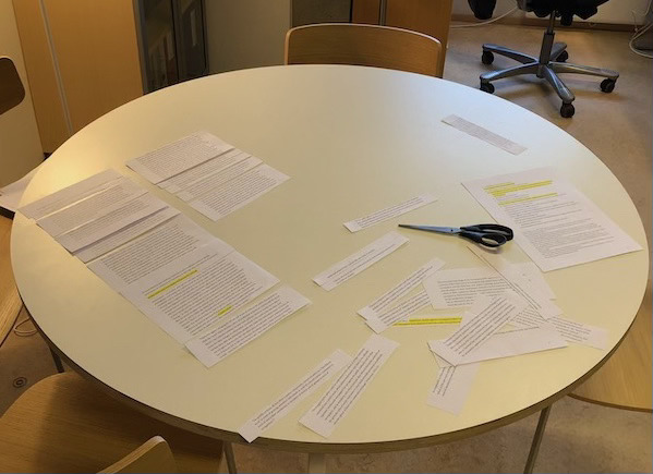 Rundt bord med en saks og en rekke oppklipte utskrifter av manussider på.